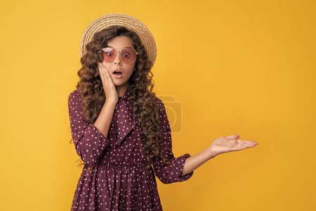 Photo for Surprised child in straw hat and sunglasses with long brunette curly hair presenting product. - Royalty Free Image