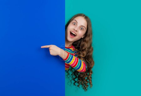 Photo for Surprised teen child behind blank blue paper with copy space for advertisement. point finger. - Royalty Free Image