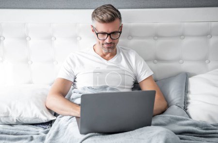 Téléchargez les photos : Mature man in glasses working on laptop in bed. digital nomad. - en image libre de droit