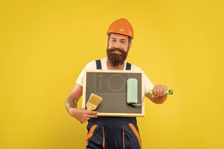 Photo for Cheerful man painter in helmet and work clothes hold paint roller brush and blackboard with copy space on yellow background. - Royalty Free Image