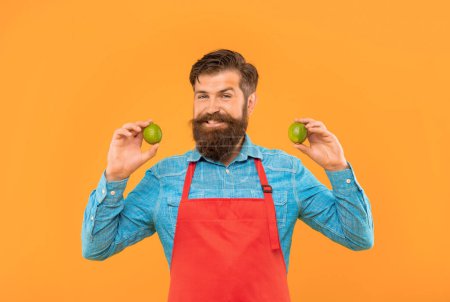 Photo for Happy man in red apron holding fresh limes citrus fruits yellow background, fruiterer. - Royalty Free Image
