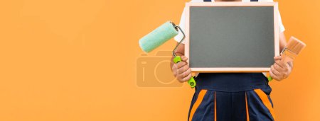 Photo for Cropped man decorator in work clothes hold paint roller brush and blackboard with copy space on yellow background. - Royalty Free Image