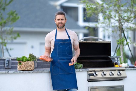 Photo for Positive man cook salmon outdoor. man cook salmon in apron. photo of man cook salmon food. man cook salmon on grill. - Royalty Free Image