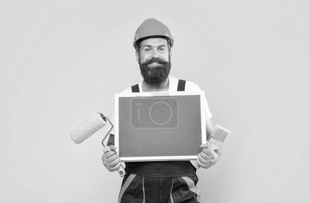 Photo for Happy man renovator in helmet and work clothes hold paint roller brush and blackboard with copy space on yellow background. - Royalty Free Image