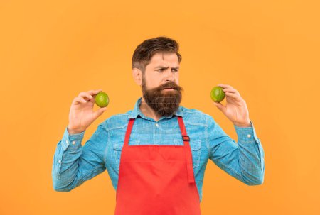 Photo for Worried man in red apron looking at fresh limes citrus fruits yellow background, fruiterer. - Royalty Free Image