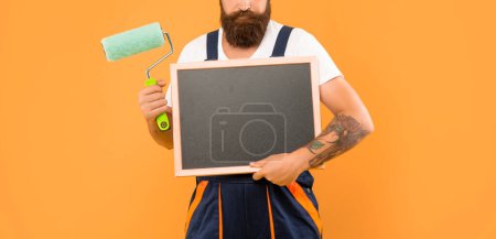 Photo for Cropped man in helmet and work clothes hold paint roller and blackboard with copy space on yellow background. - Royalty Free Image