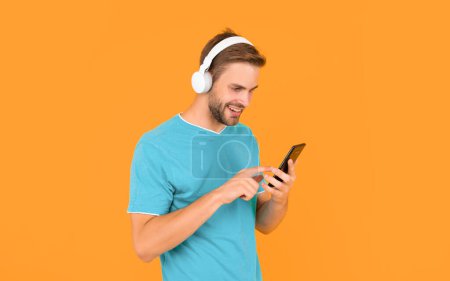 Téléchargez les photos : Joyeux jeune homme texto message sur téléphone mobile en écoutant de la musique numérique dans un casque fond jaune, sms. - en image libre de droit