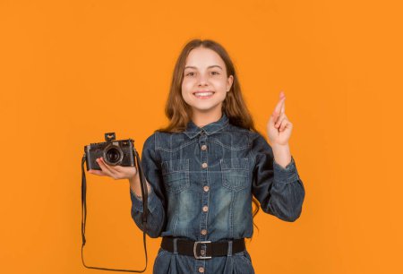 Happy superstitious girl photographer holding photo camera keeping fingers crossed yellow background, luck.