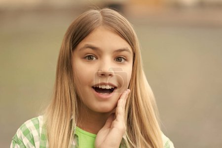 Téléchargez les photos : Portrait of tween girl with surprised face holding hand on cheek blurry outdoors. - en image libre de droit