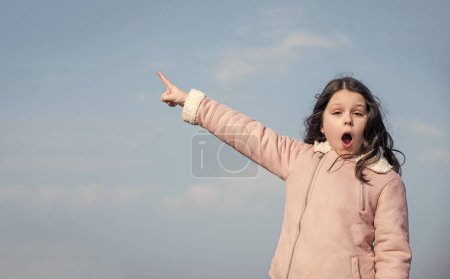 Téléchargez les photos : Enfant avec doigt de pointe du visage étonné sur fond de ciel. - en image libre de droit