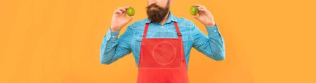 Photo for Serious man crop view in red apron holding fresh limes citrus fruits yellow background, fruiterer. - Royalty Free Image