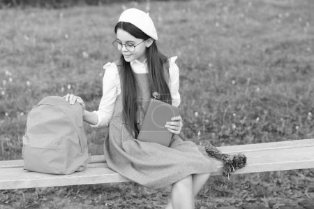 Téléchargez les photos : Enfant heureux dans des lunettes se détendre sur le banc du parc avec livre et sac à dos. - en image libre de droit