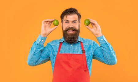 Foto de Hombre feliz en delantal rojo sosteniendo limas frescas cítricos frutas fondo amarillo, verdulero. - Imagen libre de derechos