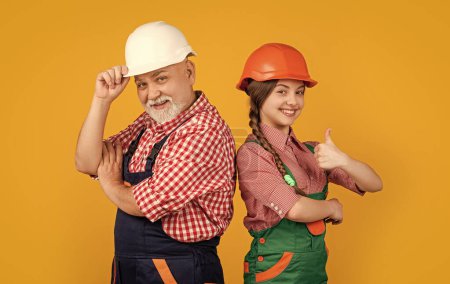 Photo for Happy teen child and grandfather builder in helmet on yellow background. thumb up. - Royalty Free Image