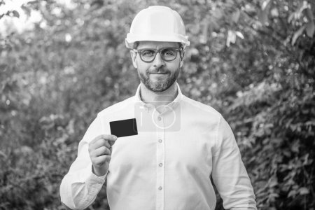 Man taskmaster in hardhat showing blank contact card outdoors, copy space.