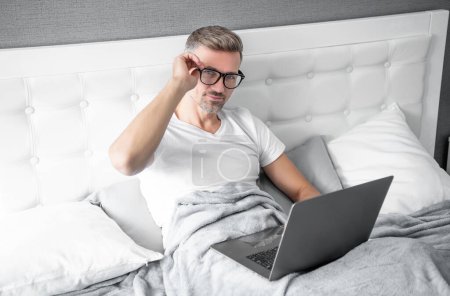 Téléchargez les photos : Mature homme dans des lunettes de travail sur ordinateur portable au lit. cyber lundi. - en image libre de droit