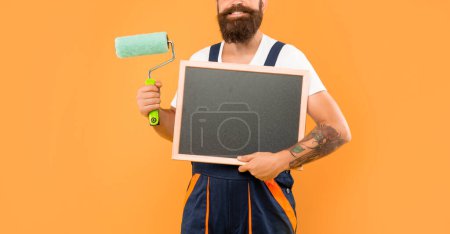 Photo for Cropped man painter in work clothes hold paint roller and blackboard with copy space on yellow background. - Royalty Free Image