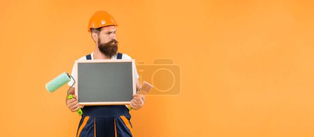 Photo for Bearded man in helmet and work clothes hold paint roller brush and blackboard with copy space on yellow background. - Royalty Free Image