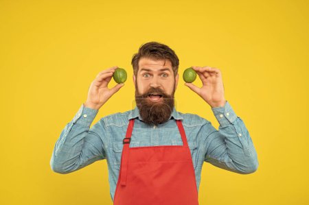 Photo for Surprised man in red apron holding fresh limes yellow background, fruit seller. - Royalty Free Image