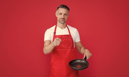 happy mature man chef in apron with frying pan on red background. point finger.