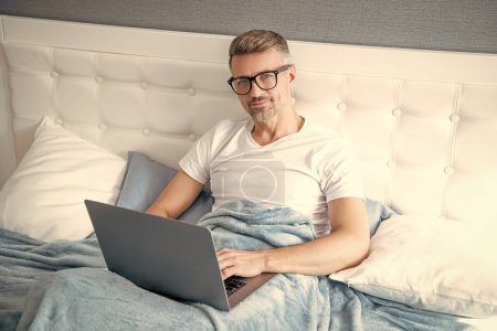 Téléchargez les photos : Mature man virtual assistant in glasses working on laptop in bed. - en image libre de droit