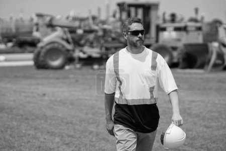 Photo for Employee construction man in vest protective hardhat walking outdoor with copy space. caucasian construction manager. construction site with heavy machine and worker. mature walking at construction. - Royalty Free Image