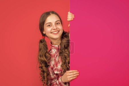 positive teen kid behind blank pink paper with copy space for advertisement.