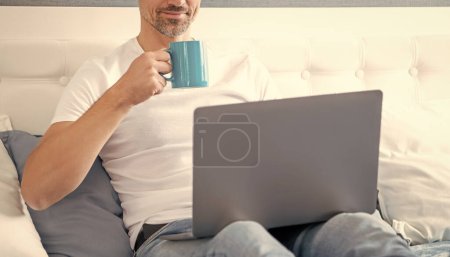 Photo for Cheerful mature man using computer in bed with coffee. - Royalty Free Image