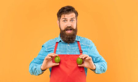 Photo for Surprised man in red apron holding fresh limes citrus fruits yellow background, greengrocer. - Royalty Free Image
