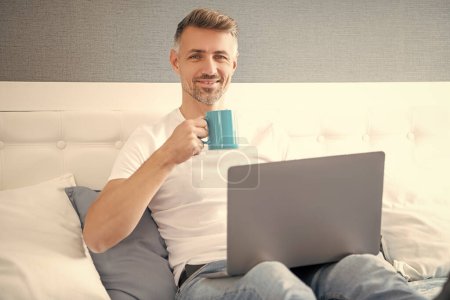Téléchargez les photos : Happy mature man using computer in bed with coffee. - en image libre de droit