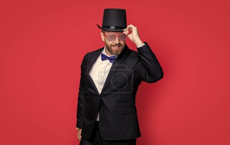 cheerful magician man wearing cylinder hat. man magician isolated on red background. magician man in cylinder hat. man magician wear tuxedo in studio.