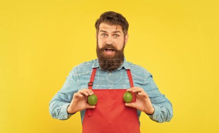 Photo for Surprised man in red apron holding fresh limes citrus fruits yellow background, greengrocer. - Royalty Free Image