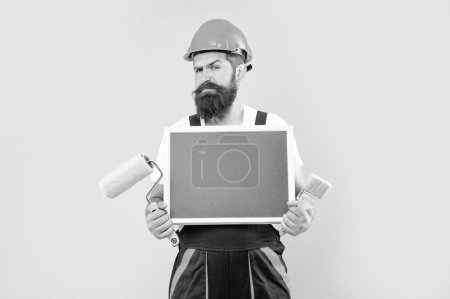 Photo for Man painter in helmet and work clothes hold paint roller brush and blackboard with copy space on yellow background. - Royalty Free Image