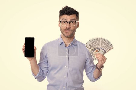 Zweifelhafter Typ mit Bargeld und Smartphone. Guy fühlt sich unsicher, ob er bar oder mit dem Smartphone bezahlen soll. Barzahlung oder Smartphone-Zahlung. Barkauf.