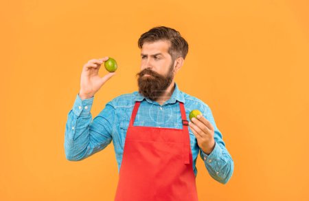 Serious man in apron looking at fresh limes citrus fruits yellow background, greengrocer.