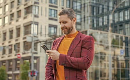 Photo for Cheerful man messaging on smartphone in the street. man messaging on smartphone outside. photo of man messaging on smartphone in jacket. man messaging on smartphone outdoor. - Royalty Free Image