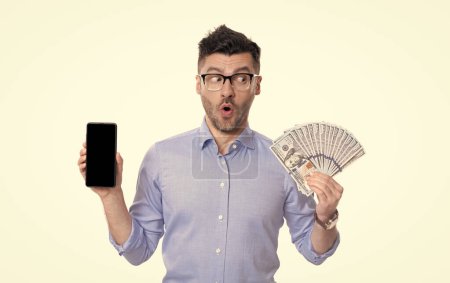 shocked guy showing cash money and smartphone. Guy feeling uncertain about paying in cash or by smartphone. Cash payment or smartphone payment. Cash purchase.