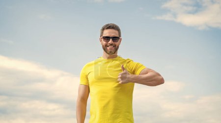 Ein fröhlicher Mann in gelbem Hemd und Sonnenbrille vor himmelblauem Hintergrund. Daumen hoch.