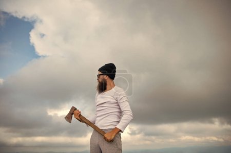 bearded man lumberjack in hat has axe. bearded man lumberjack with axe. photo of bearded man lumberjack holding axe in the mountain. bearded man lumberjack hold axe outdoor.
