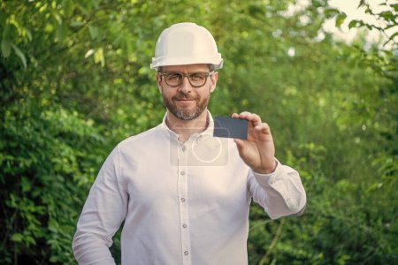 Businessman man in hardhat showing blank business card outdoors, copy space.