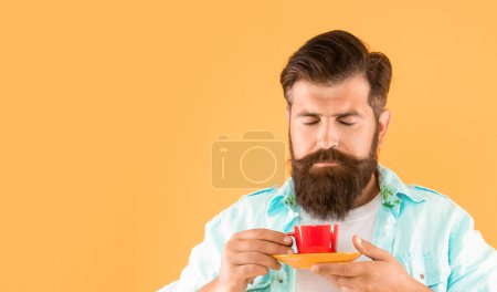 Porträt eines Mannes, der Tee riecht, mit geschlossenen Augen, Tasse und Untertasse vor gelbem Hintergrund, Kopierraum.