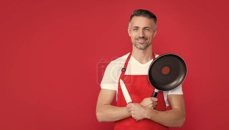 glad mature man chef in apron with frying pan and knife on red background.