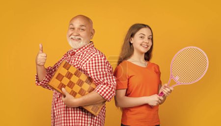 cheerful girl with grandfather with badminton racket and chess on yellow background.