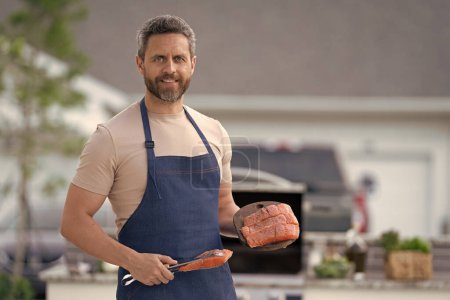 Hombre cocinar salmón en delantal, bandera. foto del hombre cocinar comida salmón. hombre cocinar salmón a la parrilla. hombre cocinar salmón al aire libre.