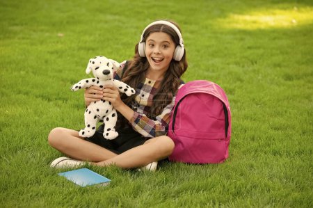 Überraschtes Teenie-Mädchen, das nach der Schule Musik hört und Hundespielzeug auf dem Gras hält. zurück zur Schule. Schüler in der Schulzeit.