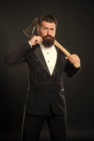 Serious bearded man twirling mustache while carrying axe at dark background, barber.