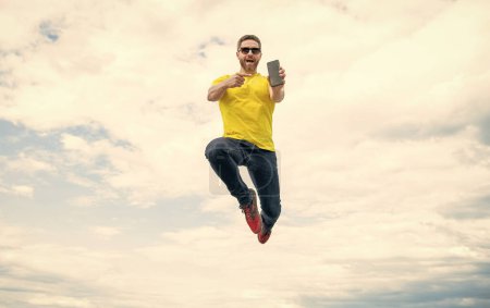 man jumping and presenting smartphone on sky background.