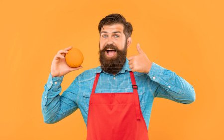 Happy man in apron giving thumb to fresh orange citrus fruit yellow background, fruiterer.