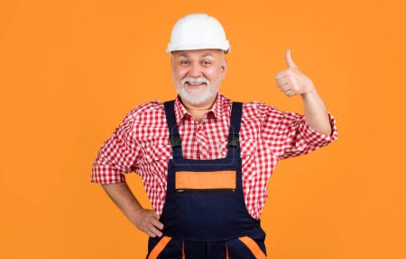 happy aged man builder in helmet on yellow background. thumb up.