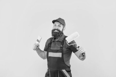 Photo for Happy bearded man painter in work clothes hold paint roller and brush on yellow background. - Royalty Free Image